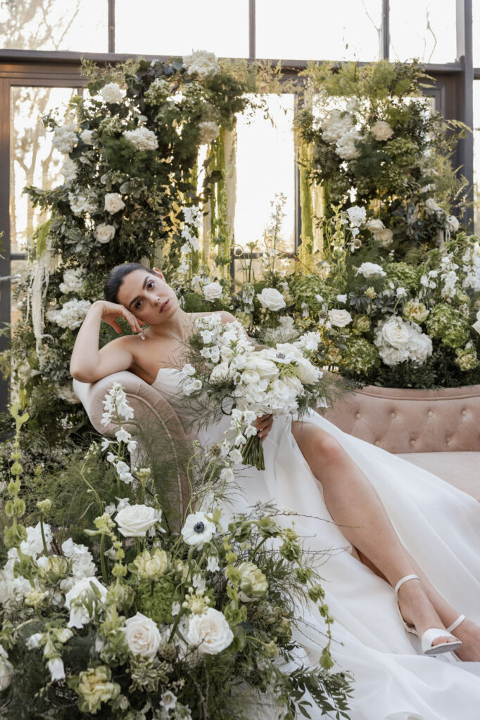 Eine Braut sitzt auf einem Sofa inmitten eines Blumenmeers und hält ihren Brautstrauß im Arm. Die üppigen floralen Arrangements umrahmen sie in weichen Farben, während ihr Kleid elegant über das Sofa fällt. Das Licht betont die Details der Blüten und des Stoffes. Die Szene wirkt romantisch, luxuriös und zeigt einen Moment, wie er typisch für exklusive Hochzeiten ist, die im Rahmen der Planung durch Sophia von Aspern und von Aspern Weddings in Hamburg, Bremen, Hannover und Niedersachsen begleitet werden.