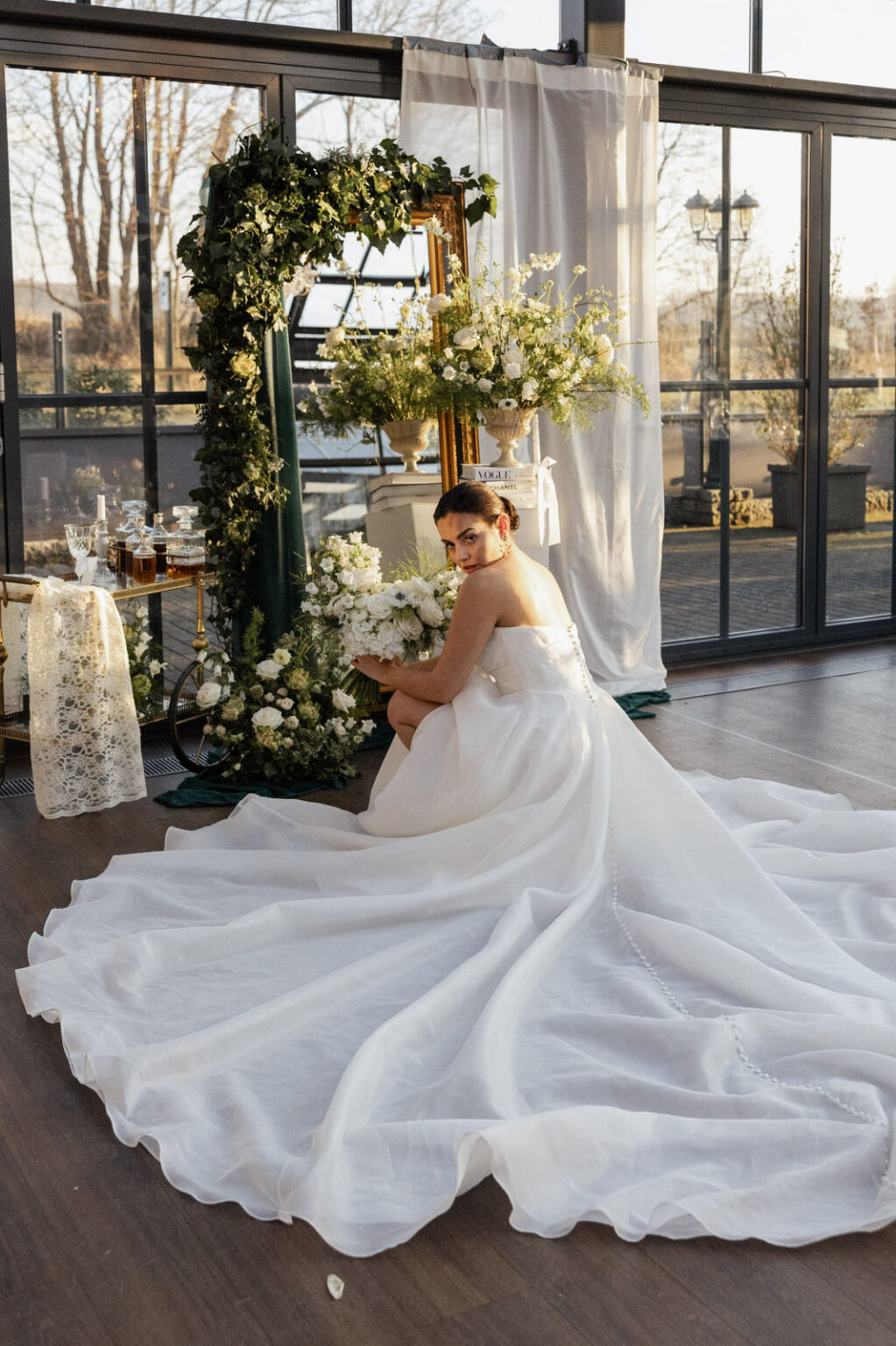 Eine Braut hockt elegant vor einem großen Spiegel und richtet ihren Blick auf ihr Spiegelbild. Ihr Kleid fällt weich um sie herum, während das Licht die Stoffdetails und ihre Konturen hervorhebt. Die Szene wirkt intim, stilvoll und modern – ein Moment, wie er oft bei Hochzeiten entsteht, die von Sophia von Aspern und von Aspern Weddings begleitet werden.