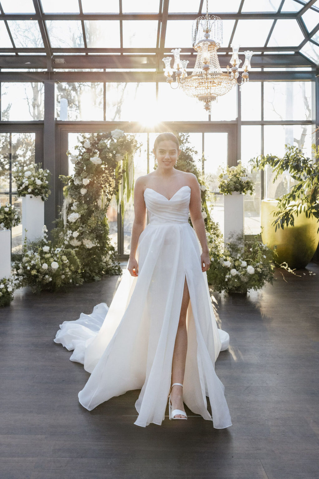 Braut im Licht vor dem Trauungssetup mit Kronleuchter Eine Braut steht vor einem Trauungssetup, über ihr hängt ein glitzernder Kronleuchter. Licht fällt von vorne auf sie und lässt Kleid sowie Blumenarrangements leuchten. Die Szene wirkt majestätisch, romantisch und edel – passend zu Hochzeiten, die von Sophia von Aspern und von Aspern Weddings begleitet werden.