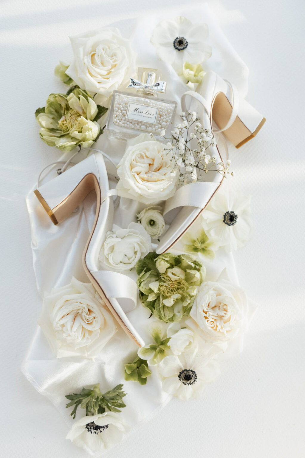 Ein Flatlay zeigt Hochzeitsschuhe, ein Parfumflakon und Blumen, sorgfältig arrangiert. Das Licht hebt die Materialien und Formen hervor und verleiht der Szene eine luxuriöse Anmutung. Dieses Bild spiegelt die liebevolle Detailplanung von Sophia von Aspern und von Aspern Weddings in der Hochzeitsplanung in Hamburg, Bremen, Hannover und Niedersachsen wider.