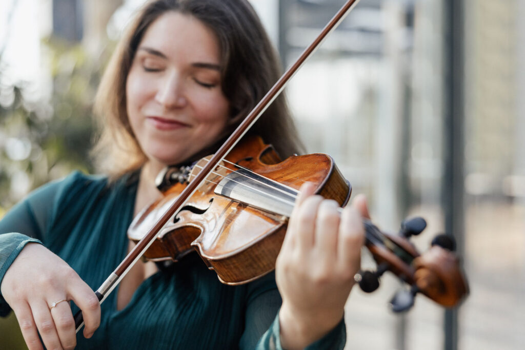 Eine Geigerin hält ihre Geige spielbereit in den Händen, der Fokus liegt auf Instrument und Körperhaltung. Der Hintergrund ist leicht unscharf, wodurch die Geige besonders hervorsticht. Die Szene zeigt die elegante musikalische Begleitung, wie sie im Rahmen der Hochzeitsplanung von Sophia von Aspern und von Aspern Weddings in Norddeutschland vorkommt.