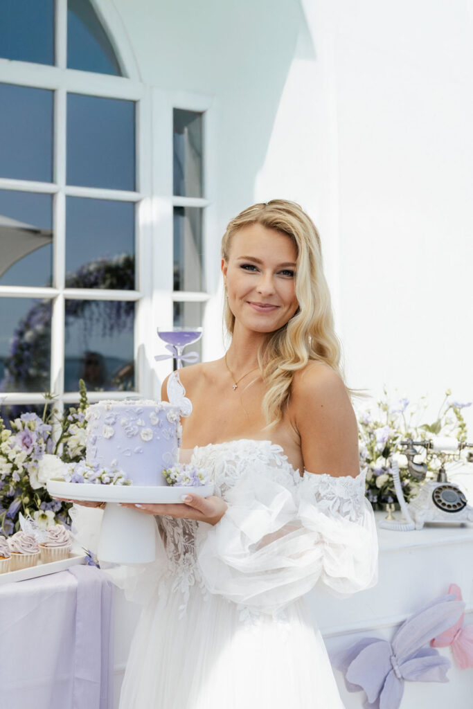 Braut mit Sonnenschirm und Glas mit lilafarbenem Drink neben der Hochzeitstorte mit violetten Cupcakes von Aura-Design-Torten im Atlantic Hotel Travemünde, elegante Hochzeitsplanung Sophia von Aspern, von Aspern Weddings, Hamburg, Bremen, Hannover, Niedersachsen.