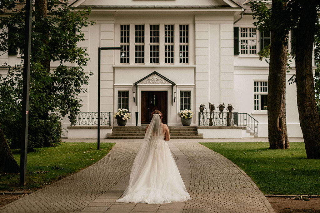 Die Braut steht vor der Villa im Heine Park in Hamburg, festlich gekleidet und strahlend – fotografiert von Kevin Kurek, Hochzeitsplanung von Aspern Weddings.