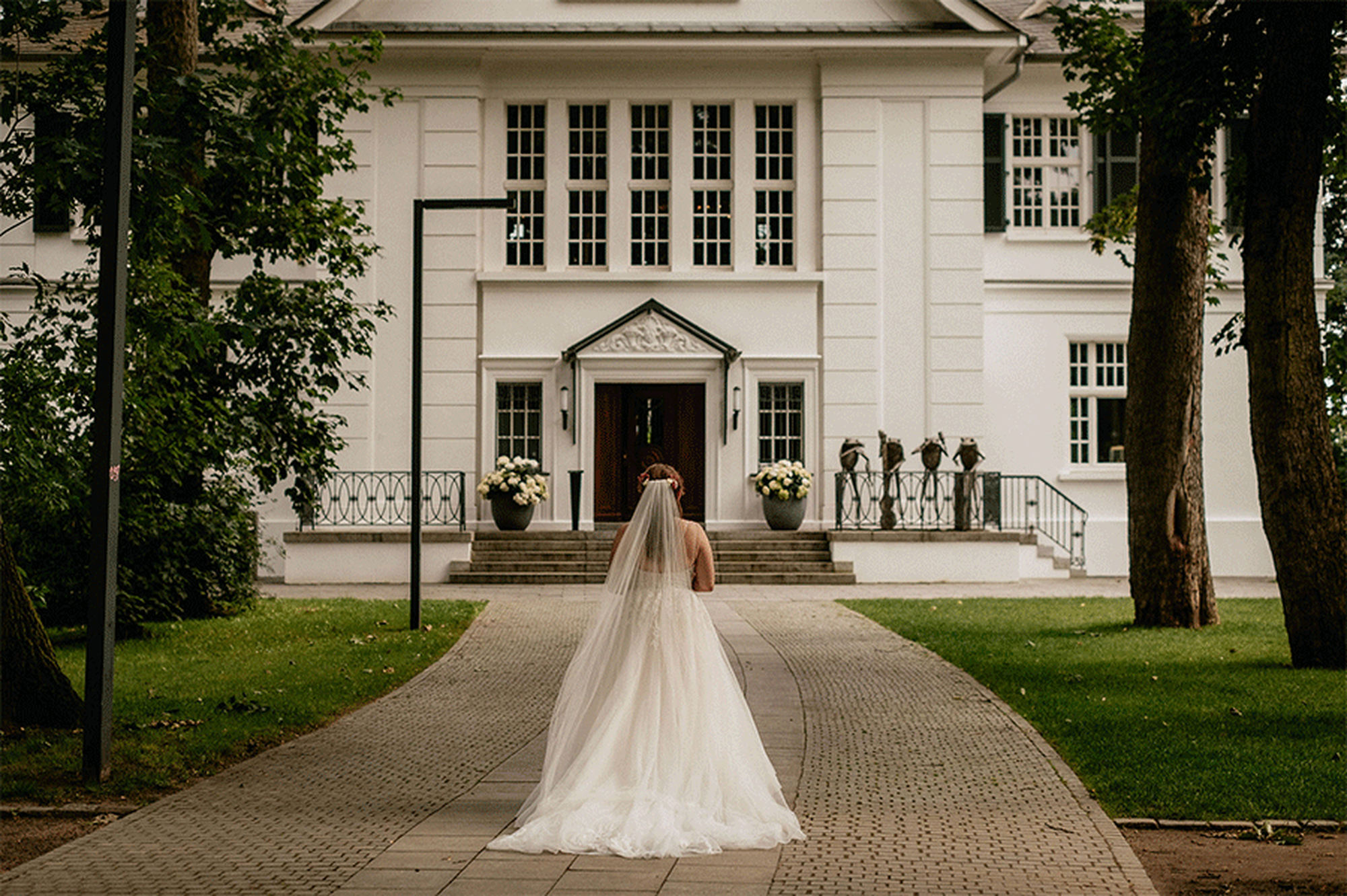 Die Braut steht vor der Villa im Heine Park in Hamburg, festlich gekleidet und strahlend – fotografiert von Kevin Kurek, Hochzeitsplanung von Aspern Weddings.