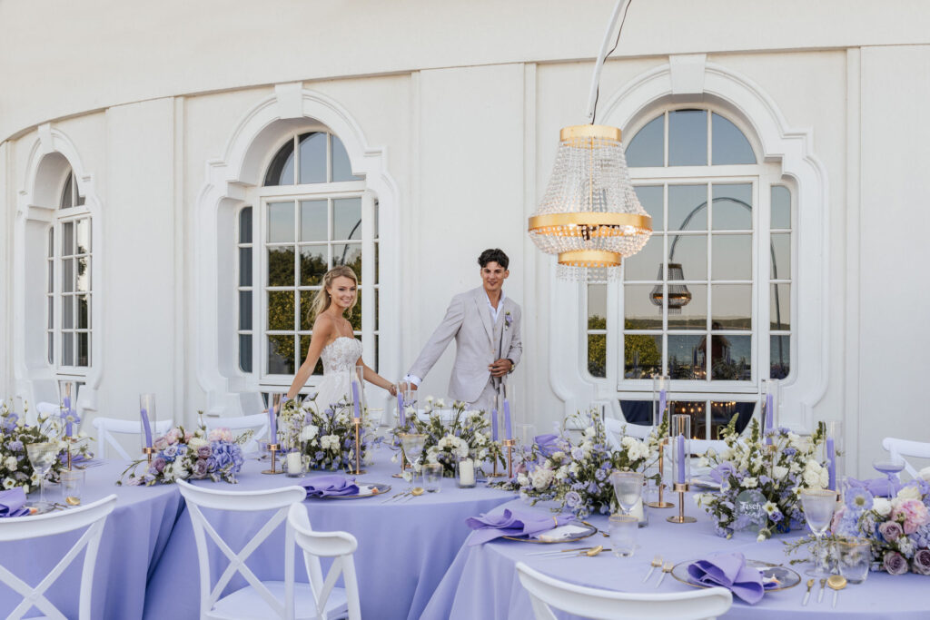 Brautpaar betrachtet das elegante Table Setting mit violetten Blumengestecken im Atlantic Hotel Travemünde, stilvolle Hochzeitsplanung Sophia von Aspern, von Aspern Weddings, Hamburg, Bremen, Hannover, Niedersachsen.