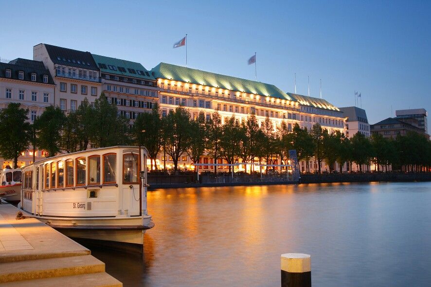 Außenansicht des Fairmont Hotel Vier Jahreszeiten in Hamburg mit Blick auf die Binnenalster. Die prächtige Fassade des traditionsreichen Hauses spiegelt sich im Wasser, während vorbeiziehende Boote und das warme Licht des Abends eine elegante Atmosphäre schaffen – ideal als Hochzeitslocation an der Elbe für exklusive Feiern in Hamburg, Bremen, Hannover und Niedersachsen.
