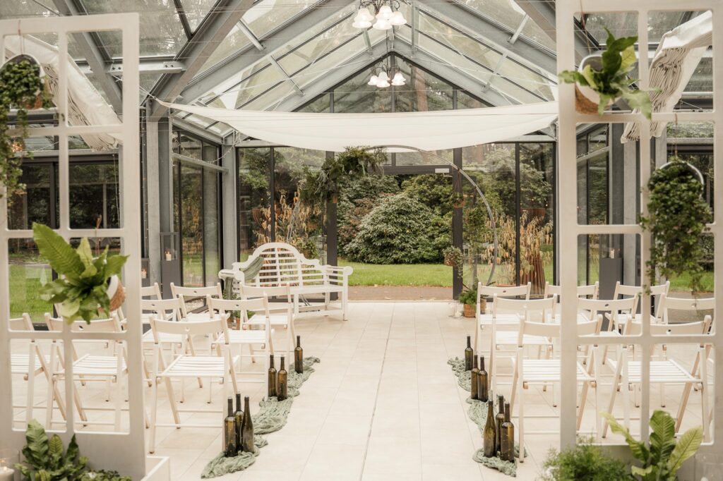Der Aufbau einer freien Trauung in der Orangerie in Niedersachsen. Zwischen hohen Glasfronten stehen weiße Stuhlreihen, die auf einen stilvoll dekorierten Traubogen aus Floristik und feinen Stoffen ausgerichtet sind. Das Licht fällt weich in den Raum und betont die elegante Atmosphäre. Sophia von Aspern als Hochzeitsplanerin richtet die letzten Details der Gestaltung aus und sorgt für ein harmonisches Gesamtbild der Hochzeitsplanung.