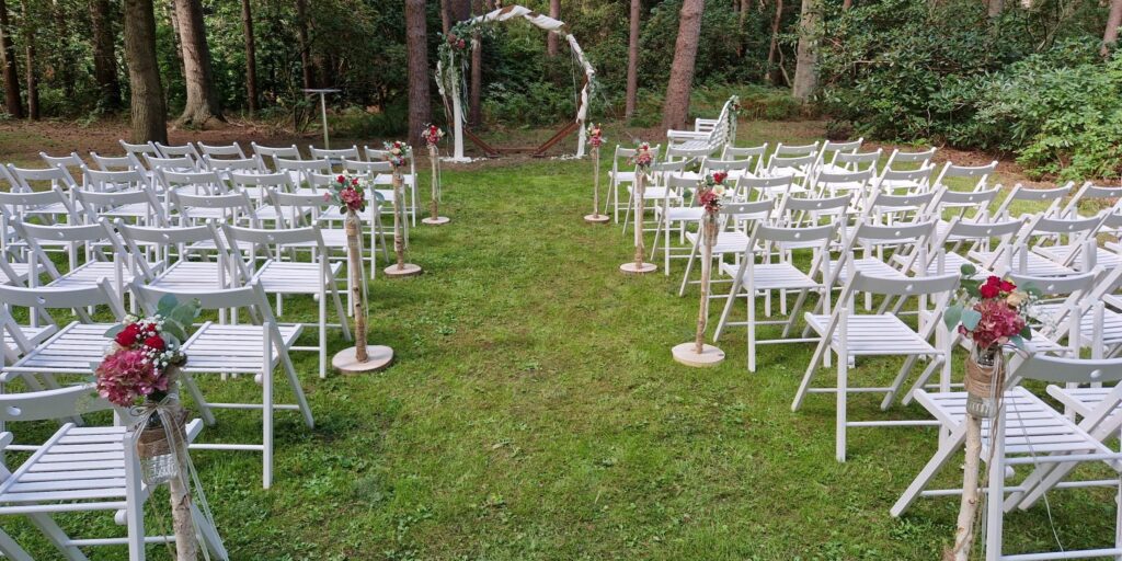 Der Aufbau einer freien Trauung in der Orangerie im Park in Niedersachsen. Weiße Stuhlreihen stehen symmetrisch vor einem eleganten Traubogen mit floraler Dekoration, umgeben von warmem Licht, Glasfronten und der klaren Architektur der Orangerie. Sophia von Aspern ist als Hochzeitsplanerin vor Ort und richtet die letzten Details des Setups aus.