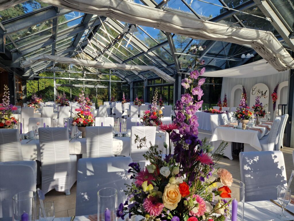 Tischdekoration in der Orangerie im Park, einer Hochzeitslocation in Niedersachsen. Auf einem festlich eingedeckten Tisch steht ein elegantes Blumengesteck in harmonischen Farben, kombiniert mit feinem Geschirr, Gläsern und dezenten Kerzen. Das natürliche Licht der Glasfronten unterstreicht die stilvolle Atmosphäre. Sophia von Aspern sorgt als Hochzeitsplanerin für ein stimmiges Gesamtbild und eine hochwertige Gestaltung der Details.