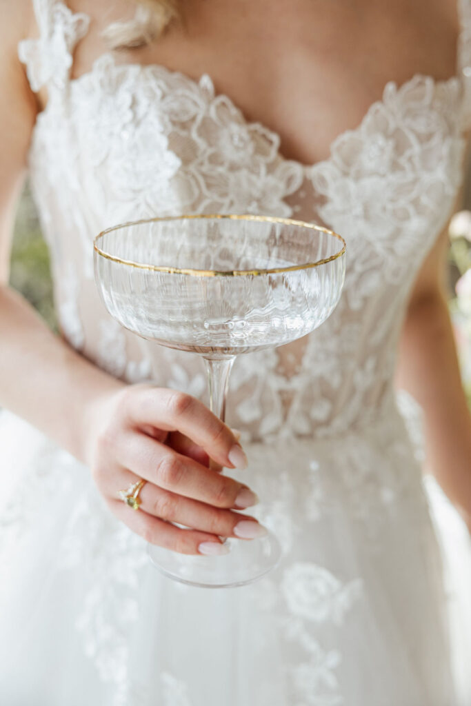 Braut hält ein Champagnerglas in der Hand, Detailaufnahme mit sichtbarem Brautkleid im Hintergrund, stilvolle Hochzeitsmomente und elegante Hochzeitsdeko bei moderner Hochzeitsplanung in Hamburg, Bremen, Hannover und Niedersachsen von Aspern Weddings Sophia von Aspern