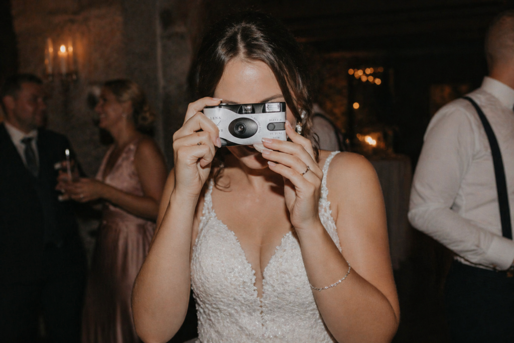 Braut hält eine Einwegkamera vor ihr Gesicht und fotografiert spielerisch während der Feier, stimmungsvolle Hochzeitsszene mit dezentem Licht und eleganter Atmosphäre, Hochzeitsplanung von Aspern Weddings in Niedersachsen, inspiriert von modernen Hochzeiten in Hamburg, Bremen und Hannover mit stilvollen Details und emotionalem Moment