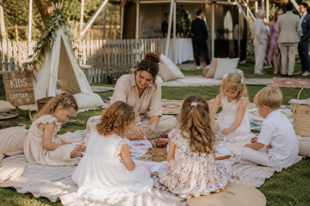 Eine Kinderbetreuung sitzt gemeinsam mit mehreren Kindern auf einer Picknickdecke bei einer Hochzeit und beschäftigt die kleinen Gäste spielerisch. Professionelle Kinderbetreuung kann bei der Hochzeitsplanung in Hamburg, Bremen, Hannover und Niedersachsen für entspannte Eltern und eine lockere Atmosphäre sorgen.