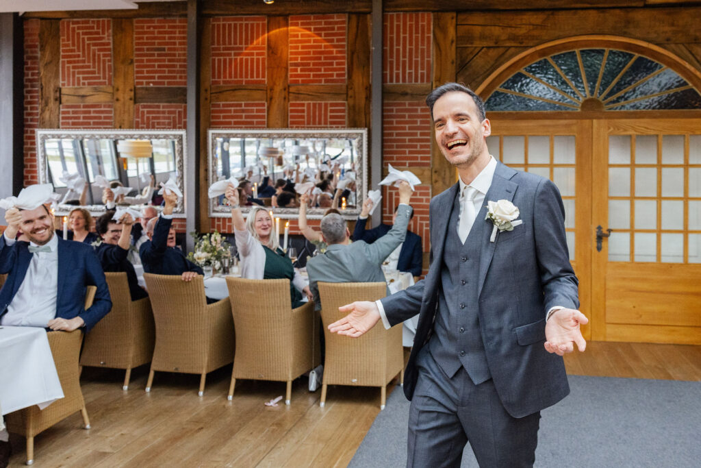 Bräutigam zieht ausgelassen in das Hotel Gut Altona ein und wedelt gemeinsam mit den Gästen mit einer Serviette. Stimmungsvoller Hochzeitseinzug als besonderer Moment einer exklusiven Hochzeit, fotografiert von Sarah Prahm und begleitet durch Hochzeitsplanung von Aspern Weddings in Hamburg, Bremen, Hannover und Niedersachsen.