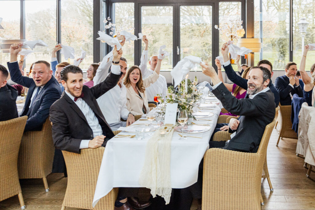 Hochzeitsgäste sitzen im Hotel Gut Altona ausgelassen an ihren Tischen und wedeln mit Servietten während der Feier. Emotionale Stimmung und besondere Party Momente bei einer exklusiven Hochzeit, fotografiert von Sarah Prahm und geplant von Aspern Weddings in Hamburg, Bremen, Hannover und Niedersachsen.