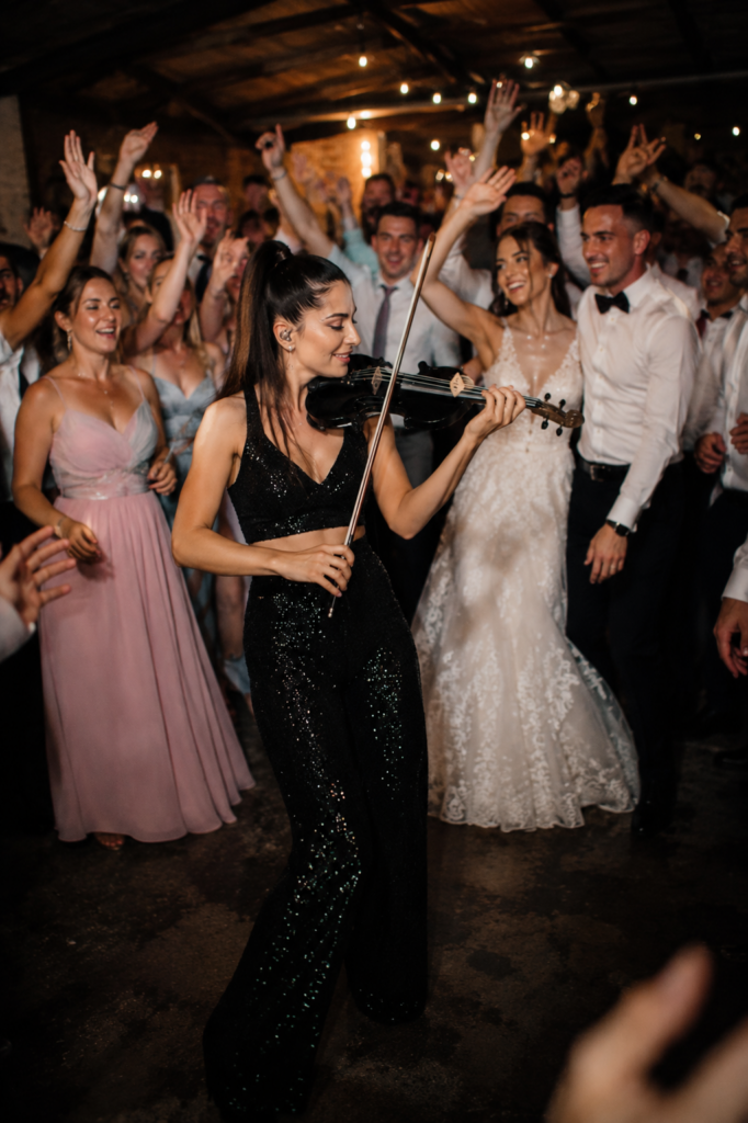 Violinistin sorgt für besondere Stimmung auf einer Hochzeitsparty, während Brautpaar und Gäste ausgelassen um sie herum feiern. Exklusive Live Musik als WOW Moment für stilvolle Hochzeiten und moderne Hochzeitsplanung von Aspern Weddings in Hamburg, Bremen, Hannover und Niedersachsen.