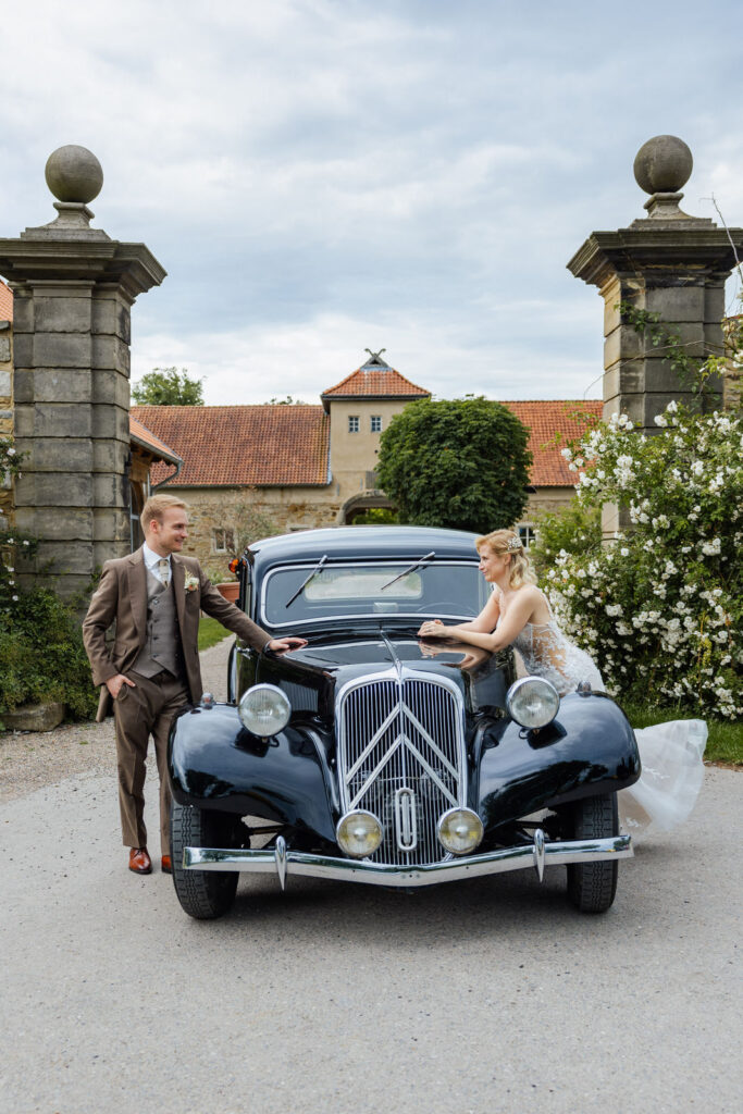 Brautpaar vor dem Torhaus des Ritterguts Remeringhausen mit Hochzeitsauto. Die historische Hochzeitslocation nahe Hannover bietet eine stilvolle Kulisse für emotionale Paarfotos. Hochzeitsplanung von Sophia von Aspern, von Aspern Weddings, in Hamburg, Hannover, Bremen und Niedersachsen.