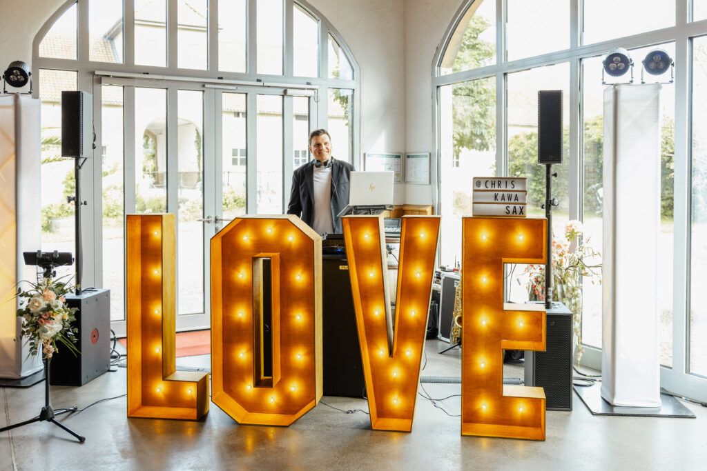 Leuchtende LOVE Buchstaben vor dem DJ im Rittergut Remeringhausen nahe Hannover. Das stimmungsvolle Setup sorgt für eine besondere Atmosphäre auf der Hochzeitsfeier. Hochzeitsplanung von Sophia von Aspern, von Aspern Weddings, in Hamburg, Hannover, Bremen und Niedersachsen. Foto: Sarah Prahm.